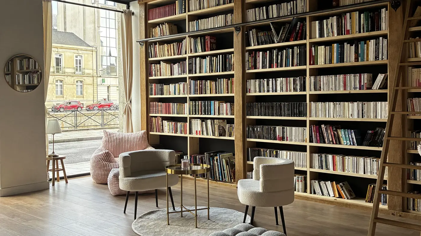 La grande bibliothèque du Quartier Littéraire à Rennes : rayonnages en bois du sol au plafond, échelle coulissante, fauteuils et vitrine ouverte sur le quai Lamennais